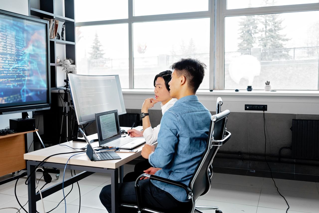 Home Two men analyzing code on computers in a modern office setting.